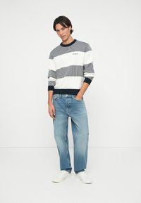 Pulover tricotat cu dungi în navy, alb și gri deschis, cu manșete și tiv ribbate; purtat cu blugi drepți albaștri deschiși și adidași albi.