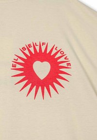 Beige t-shirt met een gedurfde rode zonnestraalprint met een hart in het midden, omringd door de tekst "SELF LOVE." Zacht katoenmateriaal.