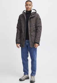 Parka gris oscuro con capucha forrada, botones a presión y múltiples bolsillos frontales. Combinada con jeans azules y zapatos de senderismo grises.
