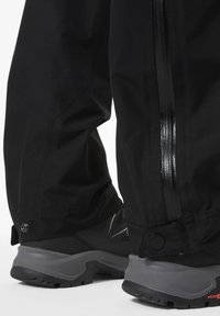 Czarne wodoodporne spodnie o eleganckim designie, z błyszczącym zamkiem po boku, w połączeniu z czarnymi butami trekkingowymi o fakturowanej podeszwie.
