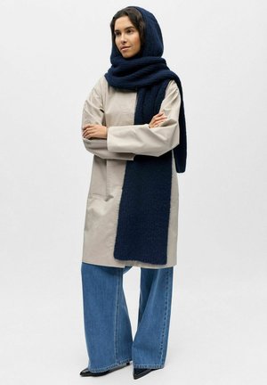 Beiger Oversize-Mantel mit langen Ärmeln, kombiniert mit einem dunkelblauen, strukturierten Schal. Locker sitzende blaue Jeans und spitze schwarze Schuhe vervollständigen den Look.