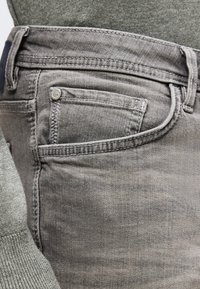 Κοντινή λήψη από τσεπάκι γκρι τζιν με εμφανή ραφές και κουμπί, φορεμένο με γκρι πλεκτό πουλόβερ.