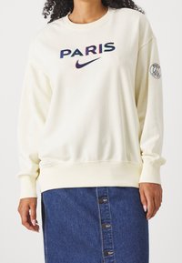 Mujer vestida con un sudadera crema que lleva la palabra "PARIS" y el logo de Nike, junto con una falda de mezclilla azul con botones, y una insignia del Paris Saint-Germain en la manga izquierda.