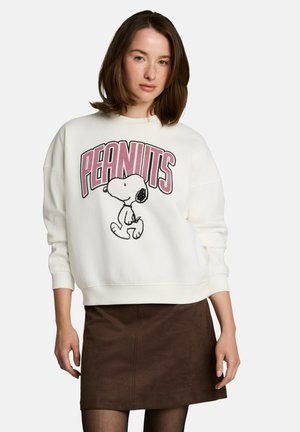 Kobieta nosząca biały sweter z różowym logo "PEANUTS" i kreskówką Snoopy'ego, zestawiony z brązową spódnicą i czarnymi rajstopami.