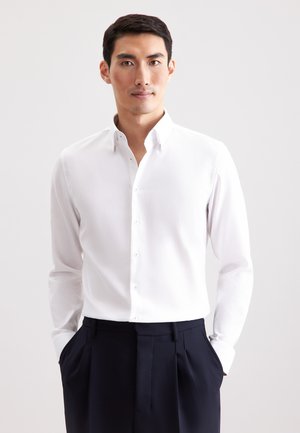 Weißes Langarmhemd mit Button-Down-Kragen, strukturierter Stoff, kombiniert mit dunkelblauen Hosen, Hände in den Taschen, neutraler Hintergrund.
