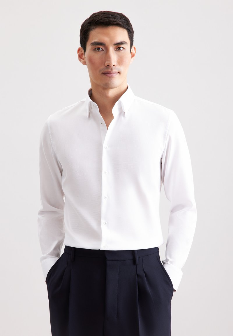 Camicia bianca a maniche lunghe con colletto button-down, tessuto strutturato, abbinata a pantaloni scuri navy, mani in tasca, sfondo neutro.
