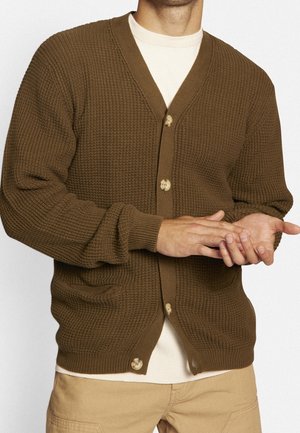 Homme portant un cardigan en maille marron avec des boutons par-dessus une chemise crème et un pantalon beige, ajustant les doigts d'une main.