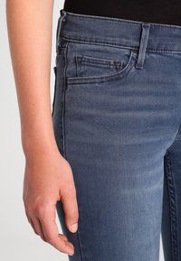 Jeans de mezclilla en un tono azul medio, con una textura suave, bolsillos delanteros y un diseño ajustado, mostrados con una mano apoyada en la pierna.