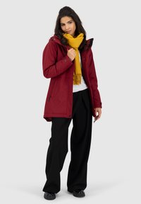Rode waterdichte jas met capuchon, gecombineerd met een gele sjaal. Model draagt zwarte wijde broek en zwarte schoenen. Eenvoudig ontwerp en strakke lijnen.