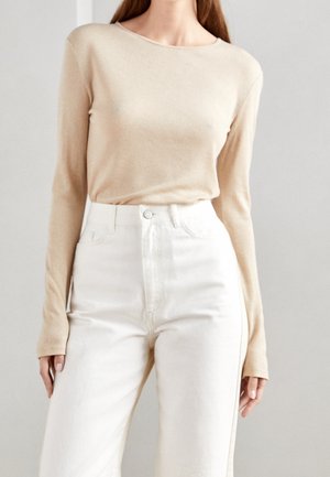Femme portant un haut beige à manches longues rentré dans un pantalon blanc taille haute, debout devant un fond clair uni.