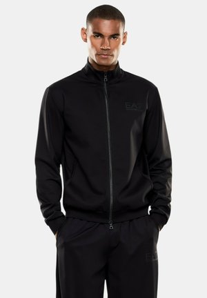 Uomo con capelli corti che indossa una giacca nera con cerniera e pantaloni neri coordinati con i logo EA7 Emporio Armani, mani nelle tasche, sfondo neutro.