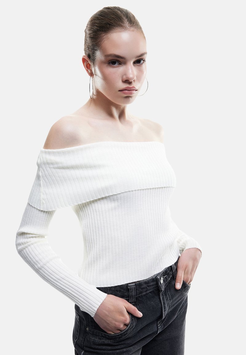 Jonge vrouw met strak naar achteren gekamd haar, gekleed in een witte off-shoulder ribbeltrui en zwarte jeans, met haar handen in de zakken, neutrale achtergrond.