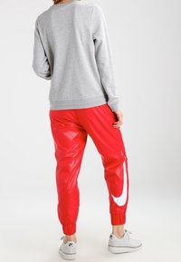 Grå sweatshirt med ribbade muddar, kombinerad med blanka röda träningsbyxor med en stor vit Nike-logga och elastiska muddar vid anklarna. Vita sneakers.