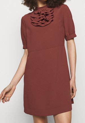 Femme portant une robe rouille à manches courtes avec des volants superposés au niveau du col montant.