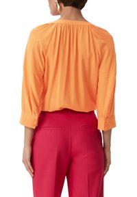Orangene Bluse mit gerafftem Rücken, Dreiviertelärmeln und geknöpften Manschetten, kombiniert mit rosa Hosen mit Gesäßtaschen.