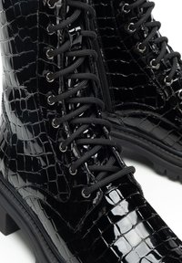 Černé lesklé kotníkové boty s krokodýlím vzorem, černými tkaničkami, bočním zipem a masivním podpatkem na bílém pozadí.