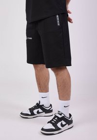 Pantalones cortos de algodón negros con un bolsillo lateral, con el detalle de texto en blanco "XX PARIS" y complementados con zapatillas Nike en negro y blanco y calcetines tipo crew.