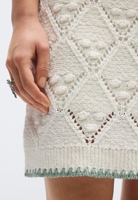 Heledad valge kudumkleit tekstuurse teemantmustriga, millel on laineline roheline ääreosa. Käsi on paigutatud, et näidata detaili ja tekstuuri.