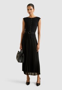 Naine kannab ilma varrukateta musta vööga pluusi, millel on nööbid, ja pleeditud musta midi-seelikut, hoidmas musta kootud käekotti.