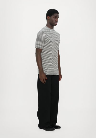 T-shirt gris à motifs avec des manches courtes et une coupe décontractée, associé à un pantalon noir large et des chaussures noires, sur fond blanc.