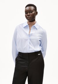 Lichtblauw overhemdsblouse met een kraag, gedragen over een bruine coltrui, gecombineerd met op maat gemaakte zwarte broeken. Glad materiaal, aangesloten ontwerp.