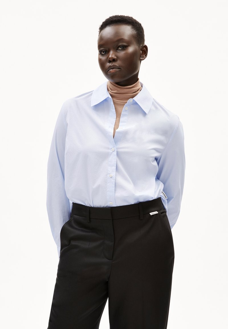 Lichtblauw overhemdsblouse met een kraag, gedragen over een bruine coltrui, gecombineerd met op maat gemaakte zwarte broeken. Glad materiaal, aangesloten ontwerp.