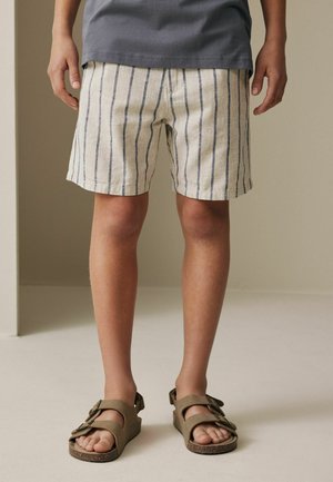 Dziecko ubrane w beżowo-granatowe paskowane krótkie spodenki, szarą koszulkę i brązowe sandały z podwójną klamrą, stojące na jasnej podłodze.