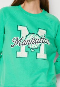 Mujer vistiendo un sudadera verde con una gran "M" blanca y el texto y emblema de "Manhattan" en la parte central del pecho.