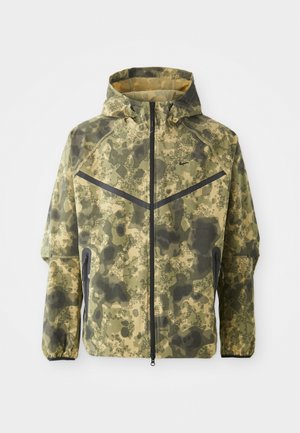 Kamerinis žalias ir rudas striukė, turinti užtrauktuką priekyje, gobtuvą ir tekstūruotą audinį su geometriniais raštais bei juodais akcentais.