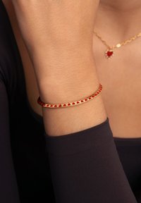 Pulsera de oro con un patrón continuo de corazones rojos a lo largo del borde exterior, llevada en una muñeca con una manga de tela oscura.