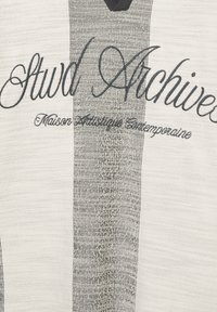 Tissu rayé gris et blanc avec une inscription en broderie foncée "Stwd Archive", présentant une surface texturée et un design en col V.