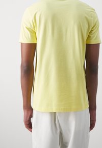 Man wearing a short-sleeve light yellow T-shirt and white pants standing with back facing the camera against a plain background.