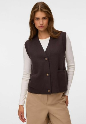 Femme portant un gilet sombre sans manches boutonné par-dessus une chemise blanche à manches longues et un pantalon beige, debout devant un fond blanc uni.
