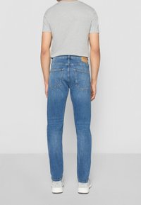 Homme portant un jean slim bleu clair, un t-shirt gris à manches courtes et des baskets blanches, debout de dos sur un fond uni.