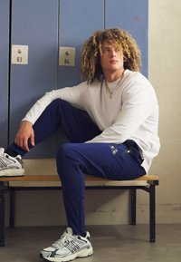 Witte long sleeve shirt, marineblauwe sportbroek en witte sneakers. Zittend op een houten bank voor grijze kluisjes.