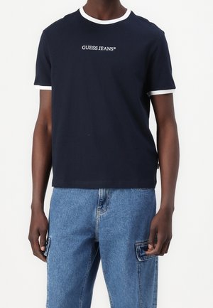 Mann trägt ein marineblaues Guess Jeans T-Shirt mit weißem Besatz an Kragen und Ärmeln, kombiniert mit blauen Denim-Cargohosen.