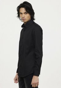 Chemise noire à boutonnières, avec un col classique, des manches longues et une coupe ajustée. Fabriquée à partir d'un tissu lisse avec un léger éclat.