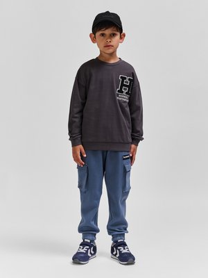 Chłopiec stojący, ubrany w czarną czapkę, ciemnoszarą bluzę z dużym logo "H", niebieskie spodnie bojówki i granatowe trampki na jednolitym szarym tle.