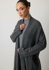 Tmavo šedý kardigán s jemnou textúrou, otvoreným predným dizajnom a dlhými rukávmi, nosený cez svetlo šedý rebrovaný top. Výrazné vrecká.