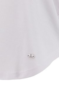 Camisa blanca texturizada con dobladillo curvado y una etiqueta ovalada plateada con el logo "KEY LARGO" en la esquina inferior derecha.