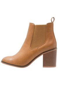 Botín tobillo marrón claro hecho de cuero con un panel elástico texturizado, puntera puntiaguda y un tacón de bloque a rayas. Incluye una pestaña para fácil colocación.