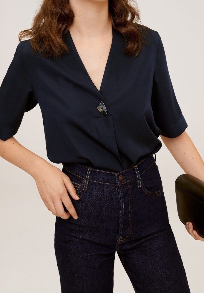 Femme portant un chemisier bleu marine avec un seul gros bouton et un jean bleu foncé taille haute, tenant une pochette noire.