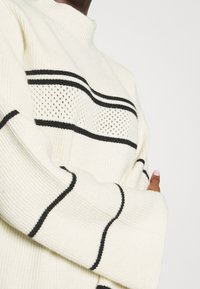 Stickad tröja i krämfärg med svarta horisontella ränder och texturerat mönster. Har hög krage och vida ärmar. Mjuk, ribbad tyg.