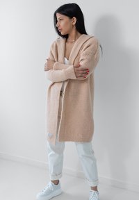 Długi, beżowy sweter z dzianiny z otwartym przodem, prążkowaną fakturą i szerokimi rękawami. W połączeniu z białymi spodniami i sneakersami.