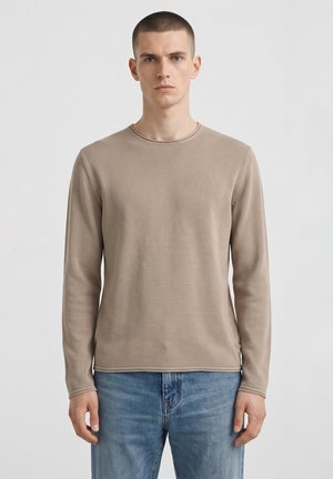 Giovane uomo con capelli corti che indossa un maglione beige a maniche lunghe e jeans blu, in piedi davanti a uno sfondo chiaro e uniforme.