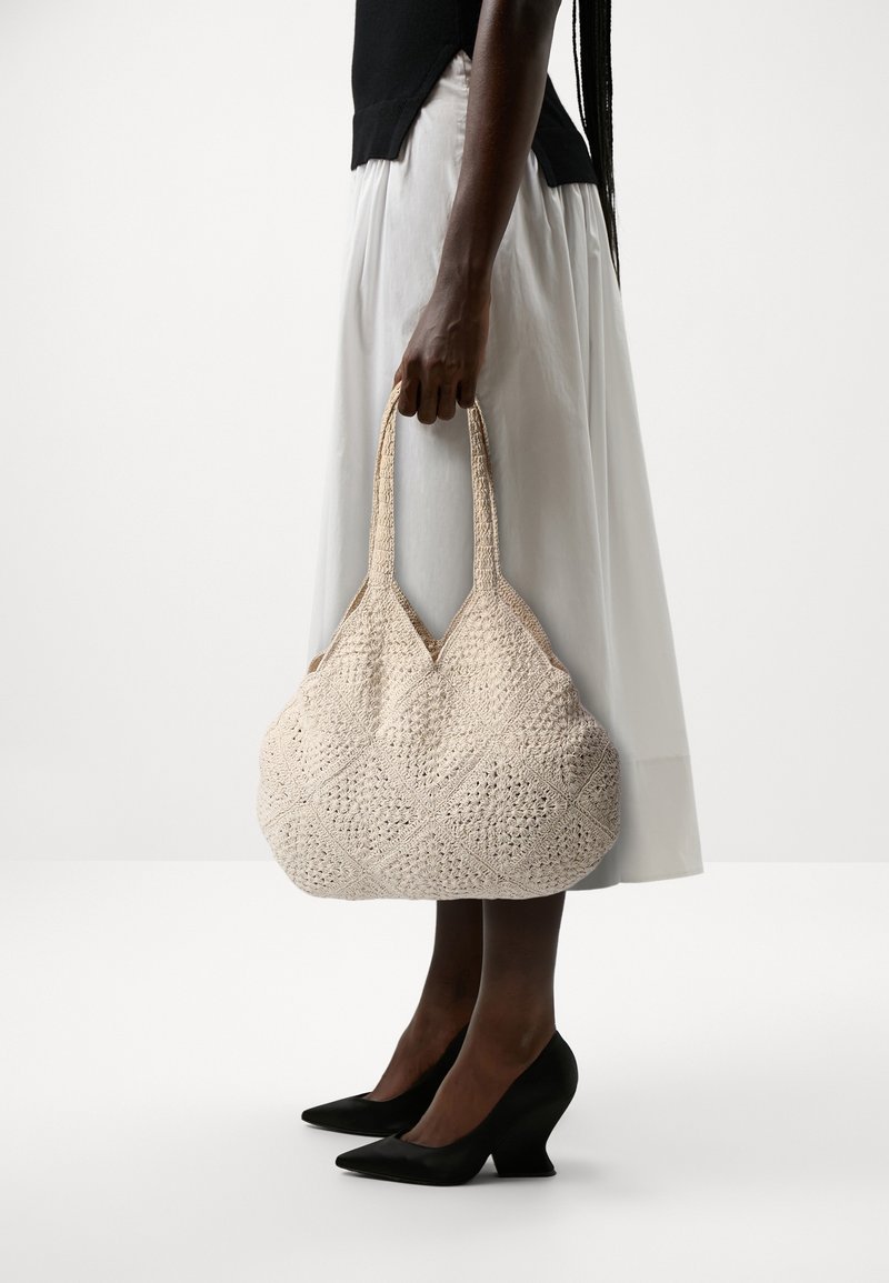 Personne portant un haut noir, une jupe midi blanche et des talons noirs, tenant un grand sac fourre-tout en crochet beige sur le côté, sur un fond uni.