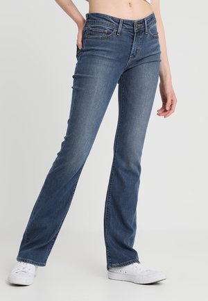 Osoba w niebieskich jeansach bootcut i białych sneakersach stoi z jedną ręką opartą na biodrze na jednolitym jasnym tle.