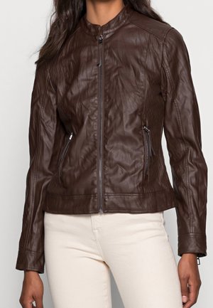 Vrouw draagt een nauwsluitend bruin leren jas met rits en twee ritszakken aan de zijkant, gecombineerd met lichtbeige broek.