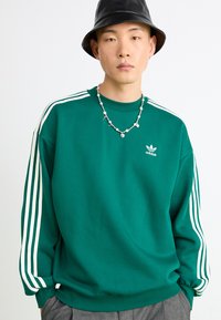 Sweatshirt verde feito de algodão com um design de três riscas brancas nas mangas, apresentando um logótipo da adidas no centro da frente. Corte casual.
