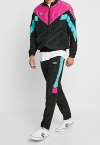 Sort træningsdragt med en zip-up jakke, der har lyserøde og turkise detaljer, kombineret med bukser, der viser lignende farvedetaljer. Hvid T-shirt under.